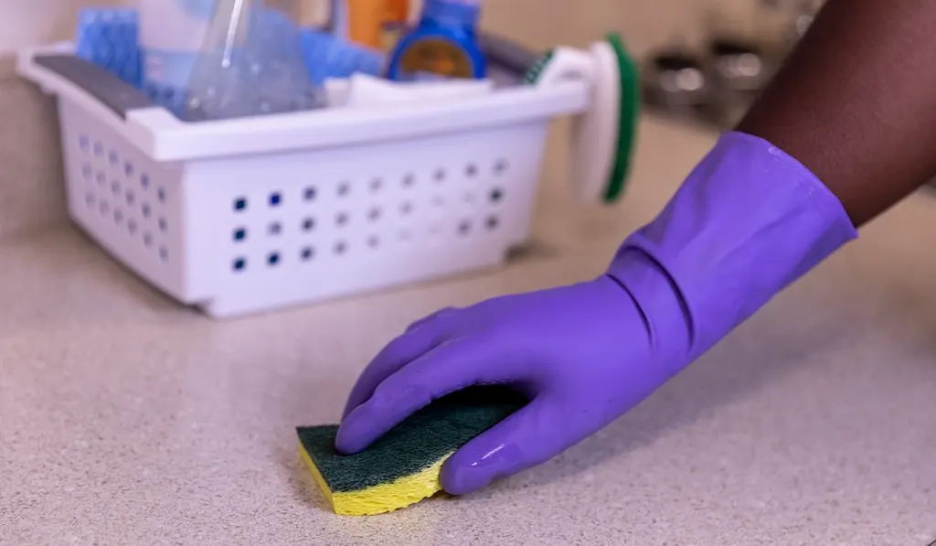 A gloved hand wiping a kitchen counter next to a cleaning caddy